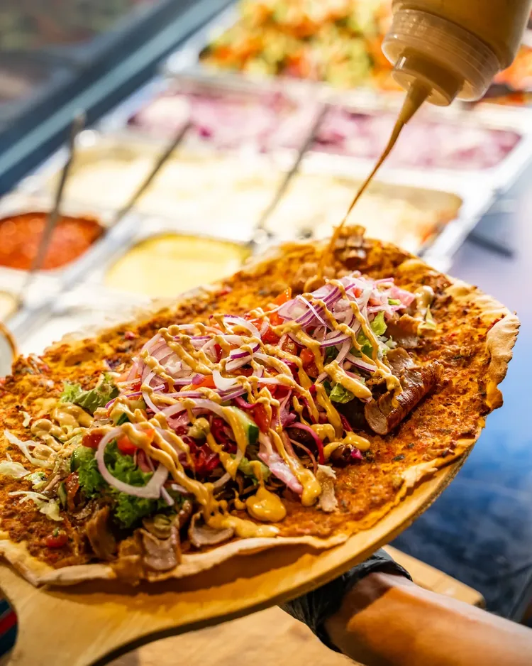 Lahmacun auf dem Holzbrett, belegt mit Fleisch, Salat und roten Zwiebeln, Sauce wird darüber gezogen